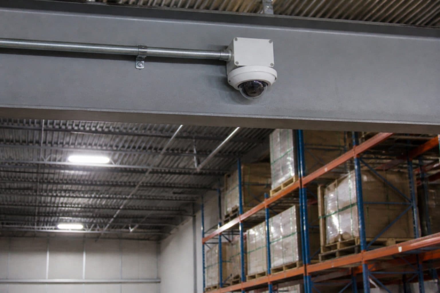 A dome security camera with its junction box and conduit on a steel beam in a warehouse